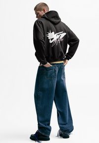 Črni pulover s kapuco z belo grafično zasnovo, kombiniran z ohlapnimi modrimi kavbojkami. Model nosi črne škornje z modrimi vezalkami.