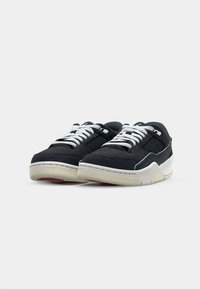 Sorte ruskinds sneakers med hvide snørebånd, meshnede paneler og cremede gummisåler, med en rød accent på ydersålen og kontrasterende syninger.