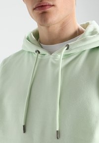 Lichtgroene hoodie met een trekkoordcapuchon, voorzien van zilverkleurige oogjes en een subtiele textuur. Gelaagd over een wit shirt.