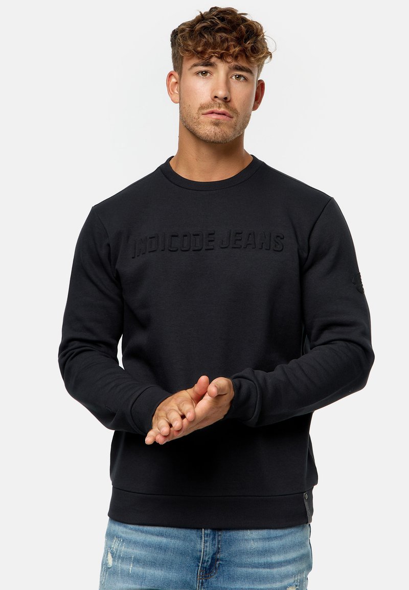 Sudadera negra con logo en relieve "INDICODE JEANS", cuello redondo, puños y dobladillo de canalé. Llevada con jeans desgastados de color azul claro.