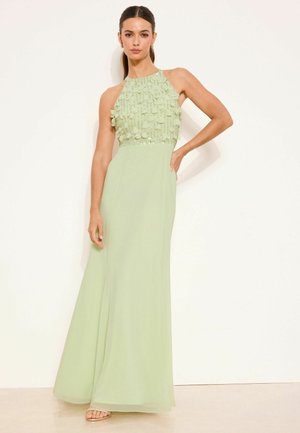 Nő pasztellzöld, ujjatlan ruhában, melynek fűzője texturált virágapplikációkkal díszített, és hosszú szoknyája van, egyik kezét a csípőjén tartva pózol.