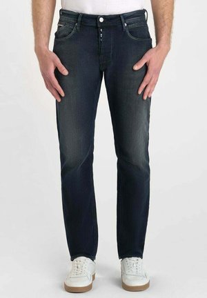 Jeans en denim bleu foncé, design à jambes droites, cinq poches, fermeture à bouton et à glissière, légères traces d'usure, portés avec des baskets blanches.