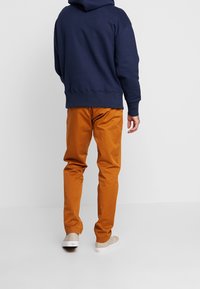 Sudadera con capucha azul marino con un bolsillo tipo canguro, combinada con pantalones chinos de color naranja. El calzado es de color beige con una ligera textura y suela clara.