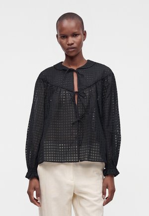 Personne portant un chemisier noir transparent à carreaux avec trois liens devant et manches longues, associé à un pantalon beige clair.