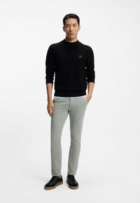 Czarny sweter o fakturze, jasnoszare chinosy oraz czarne sneakersy z brązowymi podeszwami. Model stoi prosto na jednolitym tle.