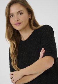 Juodas megztinis su tekstūru, trumpomis rankovėmis ir apvaliu iškirpimu. Lygus audinys su šiek tiek blizgesio ir įribotomis rankovėmis.