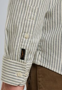 Camisa a rayas con estrechas rayas azules y crema, con puños abotonados y una etiqueta negra; la tela parece texturizada y de peso medio.