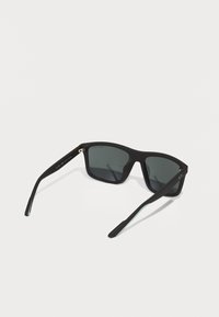 Ochelari de soare negri cu un finisaj mat. Design cu ramă pătrată, lentile întunecate și brațe subțiri cu un detaliu subtil de branding pe templu.