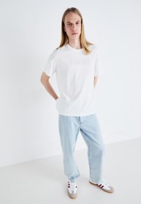 Balta trumpa rankovėmis marškinėliai su iškilniu "Levi's" logotipu, derinami su šviesiai mėlynomis laisvomis džinsomis ir pilkai baltomis sportbatėmis su raudonais akcentais.