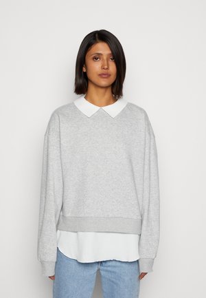 Sweatshirt gris en coton avec un col plat blanc et une taille côtelée, porté par-dessus une chemise blanche, avec des manches longues et une coupe décontractée.