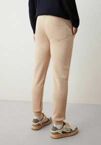 Pantaloni della tuta beige con una tasca posteriore, polsini alle caviglie e una texture liscia, abbinati a sneakers multicolori su una superficie grigia.