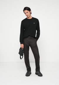 Μαύρο πουκάμισο μακρυμάνικο με πράσινο λογότυπο, καφέ υφασμένα παντελόνια, μαύρα αθλητικά παπούτσια και μικρή μαύρη τσάντα με ιμάντα. Απλός και καθημερινός σχεδιασμός.