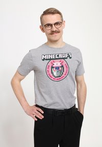 Šedé bavlněné tričko s pixel art designem, na kterém je růžový axolotl ve sklenici, orámované textem "MINECRAFT" a kulatým okrajem.