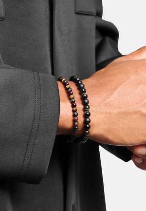 Handgelenk mit zwei Perlenarmbändern: eines mehrfarbig mit kleinen goldenen Akzenten, das andere schwarz mit matten und glänzenden Perlen, vor einer dunklen Jacke.