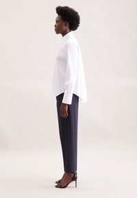 White button-up shirt with a high-low hem and long sleeves, paired with tailored navy trousers and black high-heeled sandals.