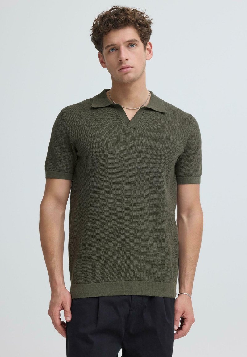 Jeune homme aux cheveux bouclés portant un polo en maille à manches courtes vert olive et un pantalon noir, debout devant un fond uni.