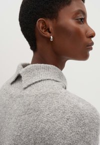 Grauer Strickpullover mit Kragen. Nahaufnahme hebt die weiche Textur und das dezente Muster hervor. Silberne Ohrringe sind an dem Modell sichtbar.