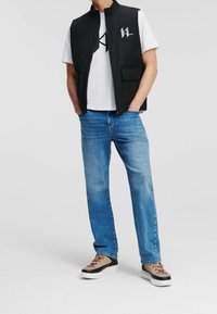 Svart ärmlös väst över en vit t-shirt med en logotyp, kombinerat med blå jeans och beige sneakers med svarta accenter och bruna snören.