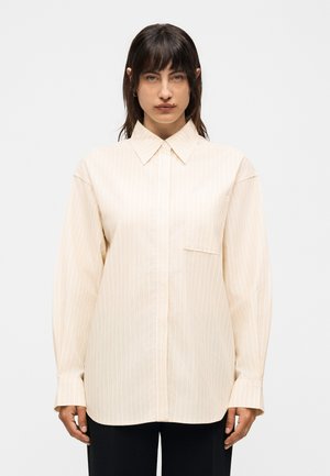 Γυναίκα με σκούρα μαλλιά που φοράει άνετο, μακρυμάνικο μπεζ ριγέ πουκάμισο με κουμπιά και μαύρο παντελόνι, στέκεται μπροστά σε απλό φόντο.