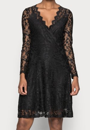 Robe noire en dentelle à manches longues avec motifs floraux, encolure en V, taille cintrée et jupe évasée longueur genou portée par une personne.