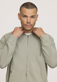 Veste à capuche zippée en tricot en tissu doux et vert clair. Elle dispose d'une fermeture éclair sur le devant, de cordons de serrage ajustables et de deux poches latérales. Texture lisse.