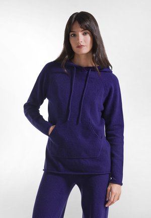 Vrouw met lang donker haar die een paarse capuchontrui en bijpassende broek draagt, staand met één hand in de zak en een neutrale uitdrukking.