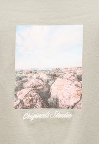 Το ανοιχτό γκρι ύφασμα διαθέτει τετράγωνη εικόνα με πετρώδες τοπίο κάτω από συννεφιασμένο ουρανό, με το κέντημα "Originals Studio" στο κάτω μέρος.