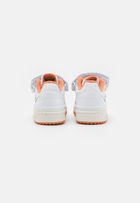 White sneakers with an orange interior, rounded heel, textured panels, and a low-top design. Hook-and-loop closure at the back.