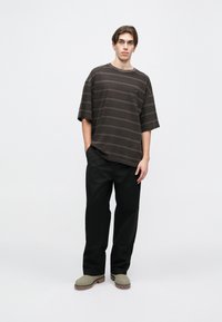 Camiseta de gran tamaño a rayas negras y marrones, de corte relajado; combinada con pantalones negros y zapatillas grises de estilo slip-on con suelas texturizadas.