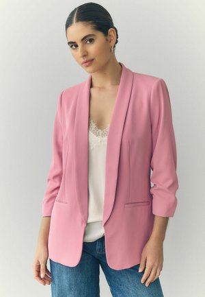 Růžový blazer se šálovým límcem, otevřenou přední částí a dvěma předními kapsami. V kombinaci s bílým topem s krajkovým lemem a modrými džínami. Hladká textura.