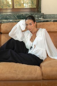Blusa bianca con volant e maniche lunghe, abbinata a una lunga gonna nera, seduta su un divano in pelle beige in una stanza ben illuminata.