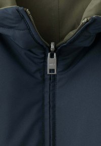 Gros plan d'une veste bleu marine avec une fermeture éclair argentée ouverte révélant une doublure intérieure vert olive et le curseur de fermeture éclair gravé MNG.
