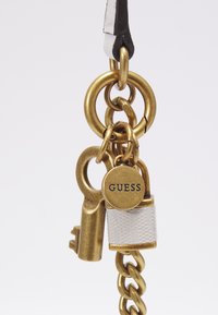 Porte-clés en laiton avec une serrure, une clé et une étiquette ronde portant l'inscription "GUESS". Comprend un métal texturé, un maillon de chaîne et un élément décoratif blanc.