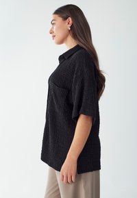 Camisa negra texturizada de manga corta, con un bolsillo y un cuello. La tela presenta un sutil patrón en relieve, combinada con pantalones beige.