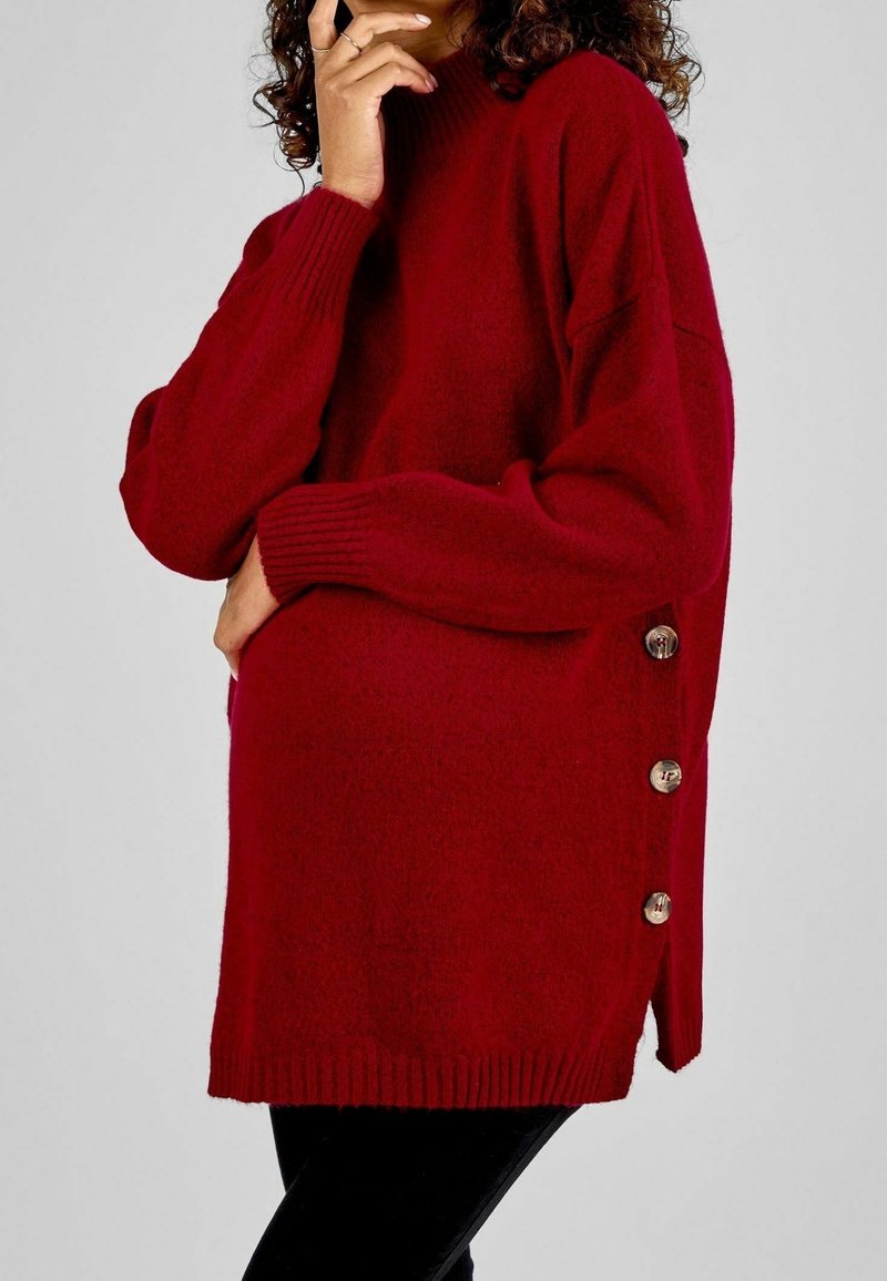 Suéter rojo de punto con cuello alto, con puños acanalados y detalles de botones laterales para un toque adicional. Material texturizado y ajuste relajado.