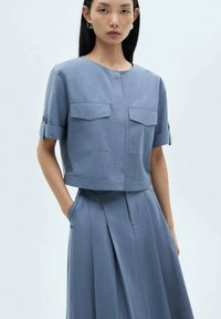 Cropped blue top with short sleeves, two chest pockets, and button accents. The fabric is smooth with a subtle sheen, paired with a matching skirt.