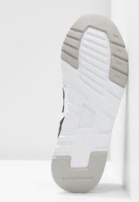 Podeszwa buta z teksturowaną, czarną powierzchnią z gumy, białą środkową warstwą i szarymi akcentami. Wyraźny wzór bieżnika zapewniający przyczepność i wsparcie.