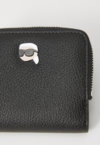 Μαύρο πορτοφόλι από υφασμάτινο δέρμα που διαθέτει ένα καρτούν φιγούρα μπίτζ με γυαλιά ηλίου, τοποθετημένο μπροστά, ενισχύοντας την λεπτομέρεια σχεδίασης.