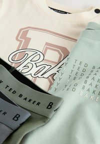 Gestapelte Kleidungsstücke umfassen ein beiges Shirt mit fettem grafischen "B" und "Baker"-Logo, grünen und grauen Stoffakzenten sowie Logodetails.