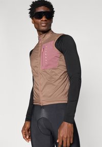 Brašna brez rokavov v rjavi barvi s pink žepom na zadrgo, iz lahkega materiala in z zadrgo spredaj. Nosi se čez črno majico z dolgimi rokavi in kolesarske kratke hlače.