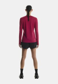 O femeie cu părul împletit stă cu fața spre spate, purtând un tricou roșu cu mânecă lungă, șorturi negre, șosete negre și pantofi sport negri.