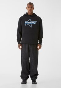 Fekete kapucnis pulóver kék "Starboy" logóval és csillag grafikával. Laza szabású, sötét farmernadrág és fekete sportcipő társaságában.