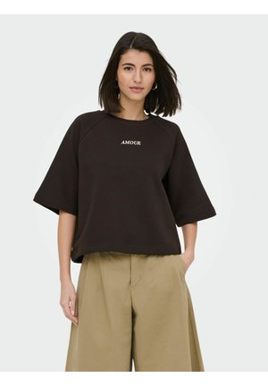 Moteris su tamsiais pečių ilgio plaukais dėvi juodą platų marškinėlį su užrašu „AMOUR“ ir smėlio spalvos platėjančias kelnes, rankos kišenėse.