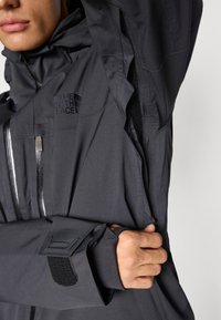 Giacca nera impermeabile con colletto alto, tasche con zip e polsini regolabili. Presenta un logo sul petto e un tessuto testurizzato.