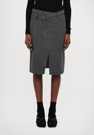 Falda vaquera en gris oscuro con una abertura frontal, cierre de botones y cintura angular distintiva. Se lleva con calcetines y zapatos negros.