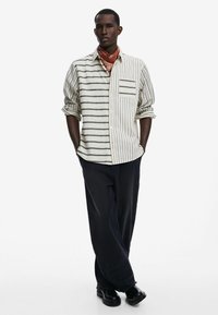 Camisa de botones a rayas en crema claro y gris oscuro, con un bolsillo en el pecho. Combinada con pantalones negros holgados y zapatos negros, y presenta una bufanda al cuello.