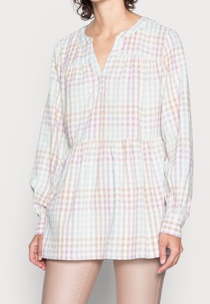 Kobieta ubrana w pastelową, kraciastą tunikę z długim rękawem, dekoltem w serek i marszczonym stanem, w połączeniu z beżowymi, dopasowanymi spodniami.