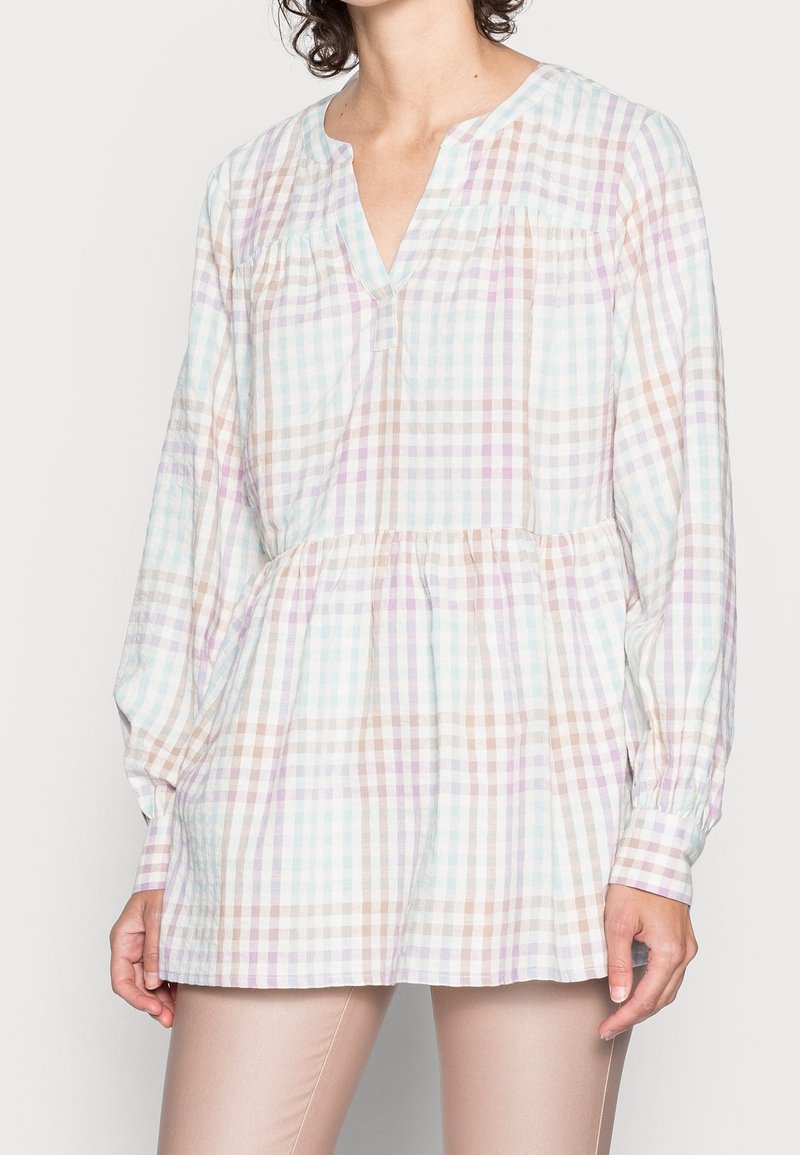 Femme portant une tunique à manches longues en tartan pastel avec un col en V et une taille froncée, associée à un pantalon slim beige.