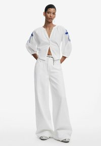 Blusa blanca corta con mangas abullonadas, bordado floral azul en los hombros, combinada con pantalones anchos blancos y zapatos plateados.