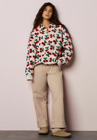 Sherpa pulover z rdečim in zelenkastim vzorcem češenj, z dodatkom zadrge do sredine vratu, kremasto ozadje. Oblečen s svetlo bež širokimi hlačami in obuvali brez vezalk.